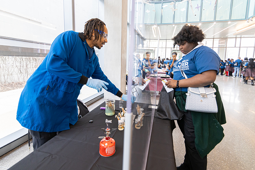 A scientist and middle school student separated by protective Plexiglas for a fire-based display at the UChicago Pritzker Molecular Engineering’s 2025 No Small Matter Molecular Engineering Fair