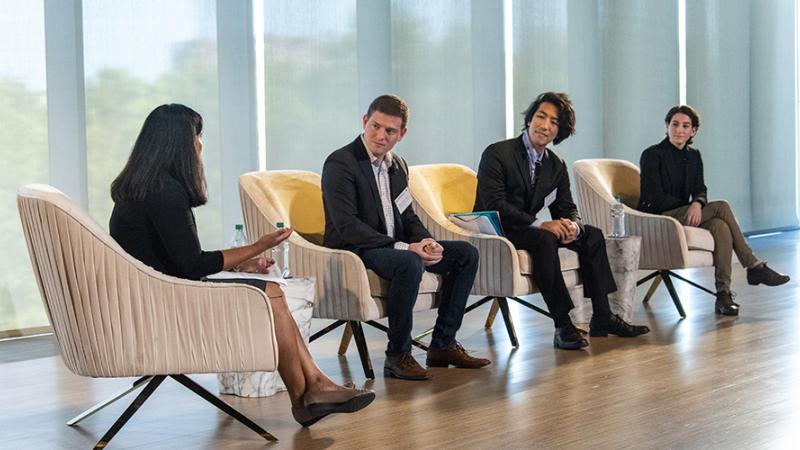 Four people are seated on a large stage, with a bank of windows behind them