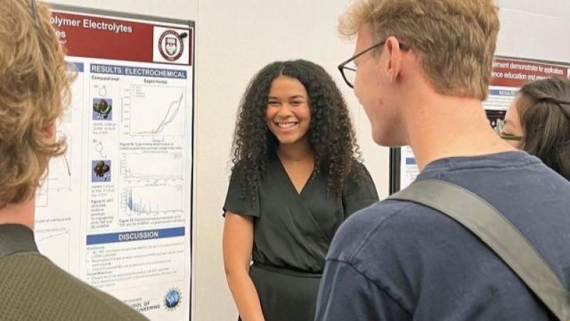 A smiling presenter stands in front of a scientific poster, speaking to a small group of listeners. Three people are seen from behind facing her, and the poster behind her includes graphs and sections like “Results” and “Discussion.”