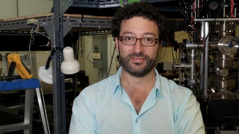 A man with dark curly hair, a beard, and glasses stands smiling with crossed arms, wearing a light blue shirt and a digital watch. He is in a lab, with complex scientific apparatus, metal racks, tubing, wires, and a large metallic chamber visible in the background.