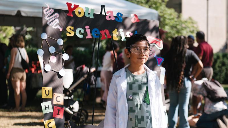 Child dressed in lab coat and goggles by a sign saying "You as a scientist" at the University of Chicago South Side Science Festival 2025