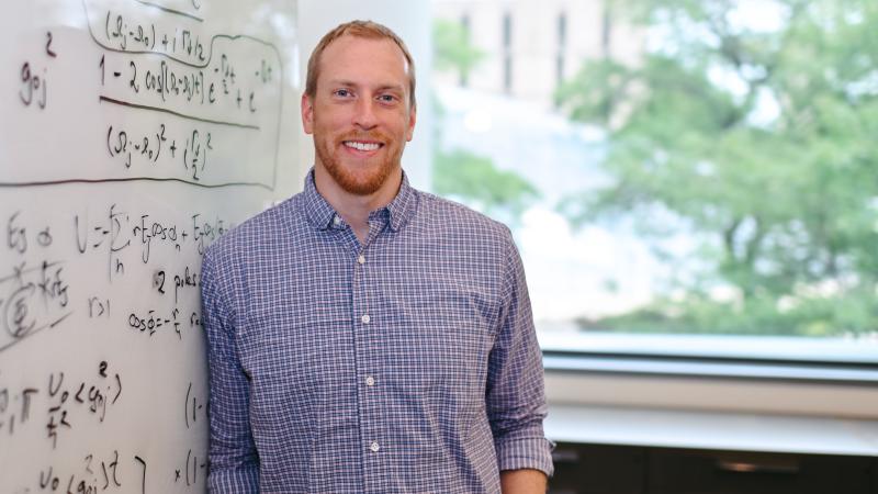 Man in button-down shirt next to whiteboard with physics equations