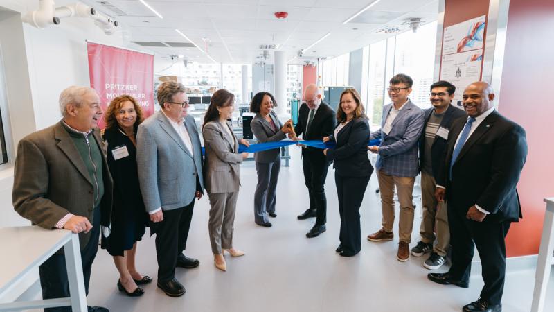 A group of nine people in professional attire celebrate a ribbon-cutting ceremony in a brightly lit modern laboratory. Two individuals in the center are cutting a blue ribbon.