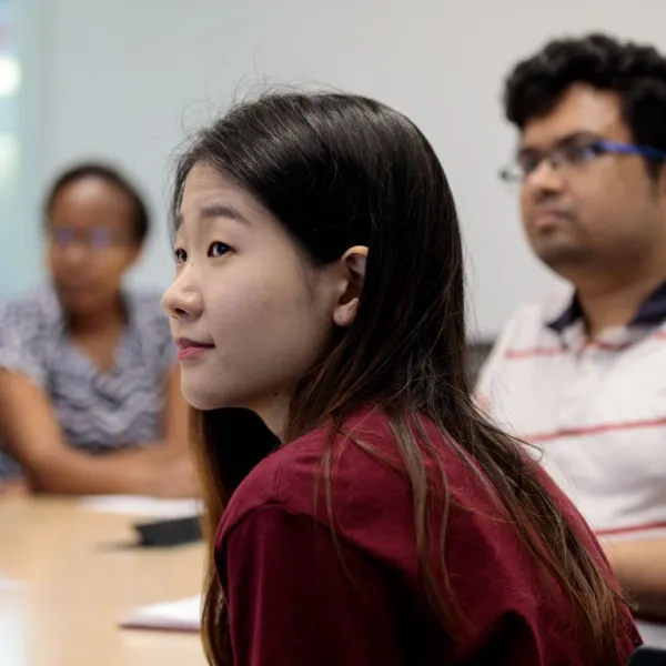 A young woman in a red shirt sits in a meeting room, turned slightly to the side and listening attentively, with two blurred colleagues seated behind her.