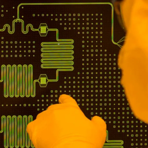 A close-up of a gloved hand pointing at a microfabricated circuit pattern with intricate lines and grids, viewed under magnification.