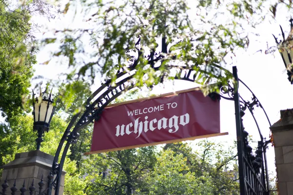 A maroon banner reading 'Welcome to UChicago' hangs from an ornate wrought-iron archway at the University of Chicago, framed by green tree branches.