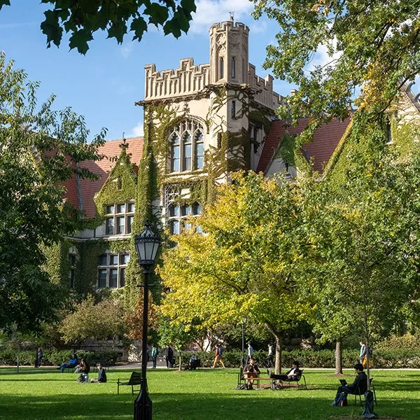 View of a Gothic-style University of Chicago building covered in ivy, framed by trees and a green lawn with students sitting and walking around campus.