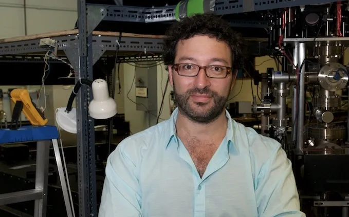 A man with dark curly hair, a beard, and glasses stands smiling with crossed arms, wearing a light blue shirt and a digital watch. He is in a lab, with complex scientific apparatus, metal racks, tubing, wires, and a large metallic chamber visible in the background.
