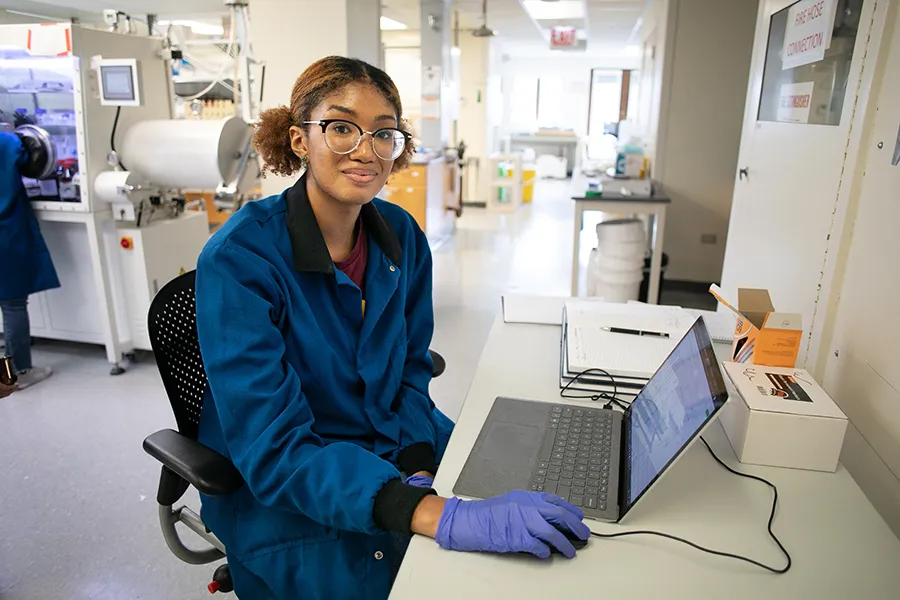 Briana Ross sits at computer with lab coat and disposable gloves