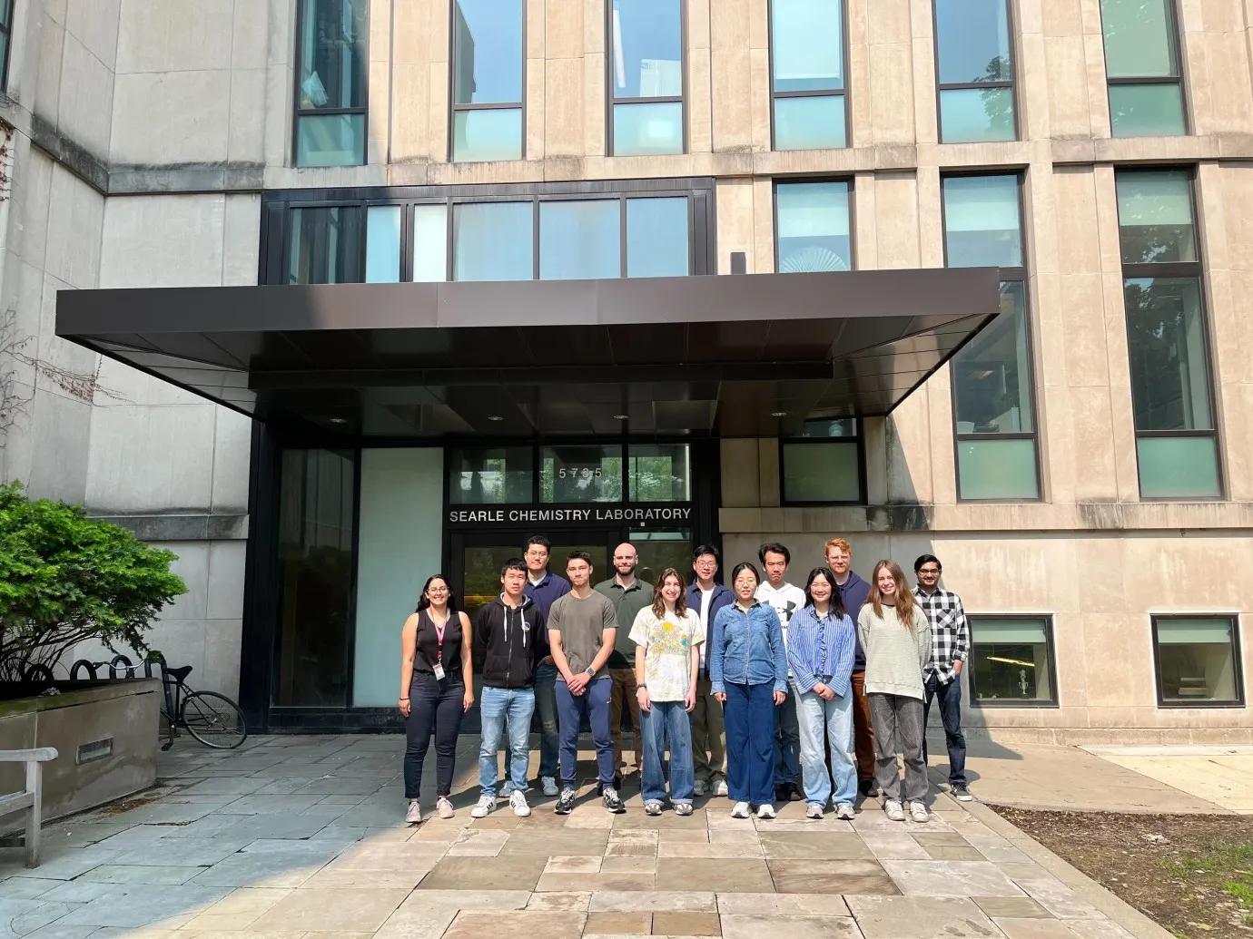 A group of about 13 people stand together outside the entrance of a modern building with large windows. Above the glass doors, a sign reads “SEARLE CHEMISTRY LABORATORY.” The group faces the camera, posing casually on a paved walkway, with the building façade forming a symmetrical backdrop.