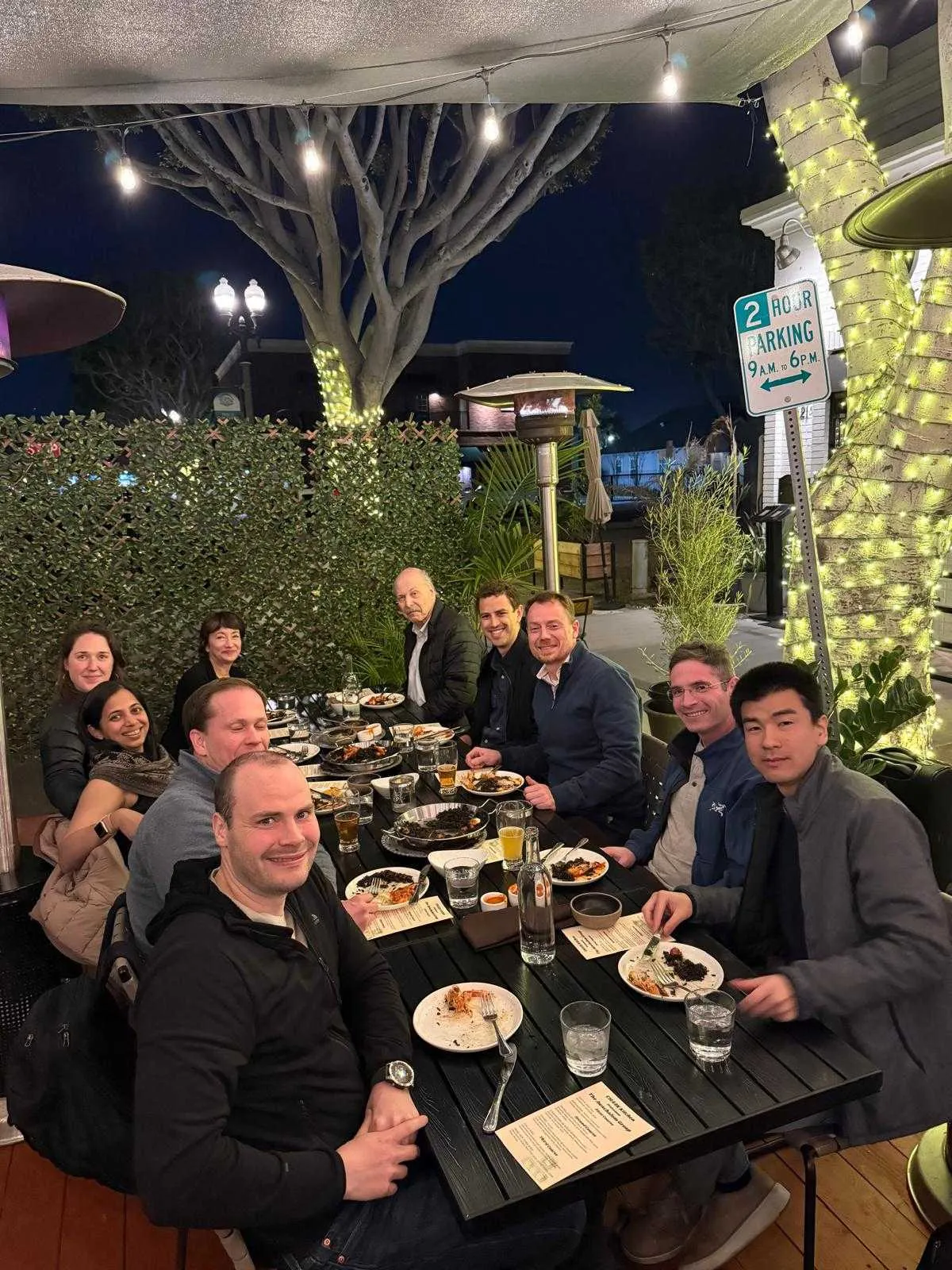 Researchers and faculty sharing a group dinner on an outdoor restaurant patio illuminated with string lights.