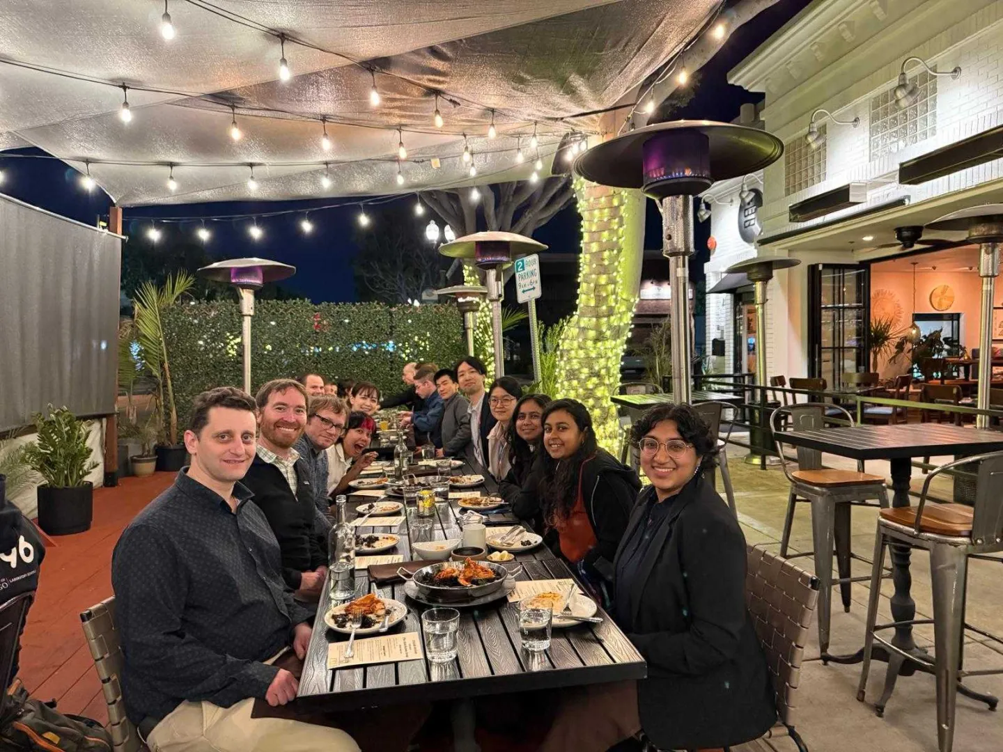 Research group dining together outdoors at night under string lights and patio heaters at a restaurant.