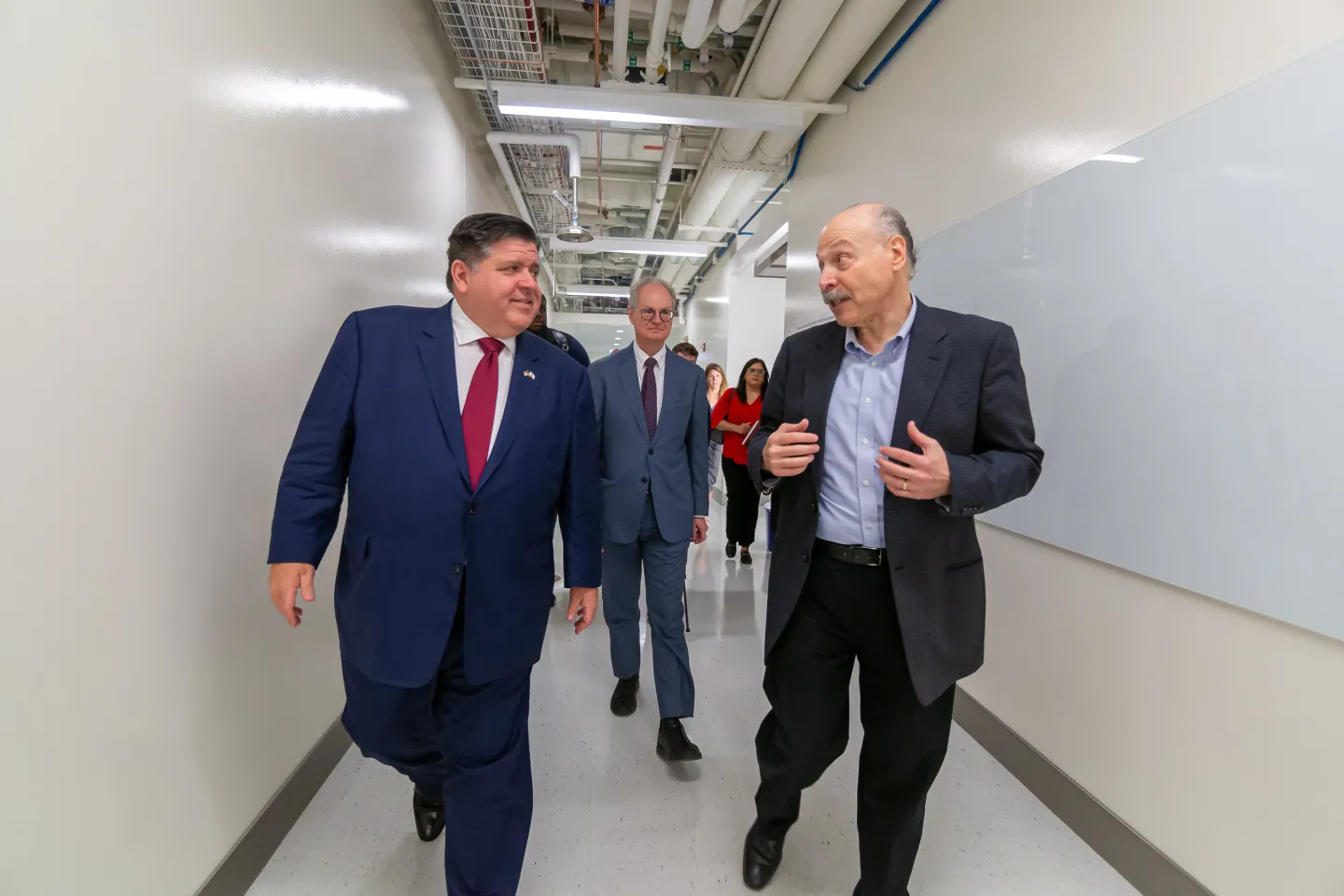 Illinois Governor J.B. Pritzker touring a research facility with David Awschalom and faculty members in a laboratory hallway.