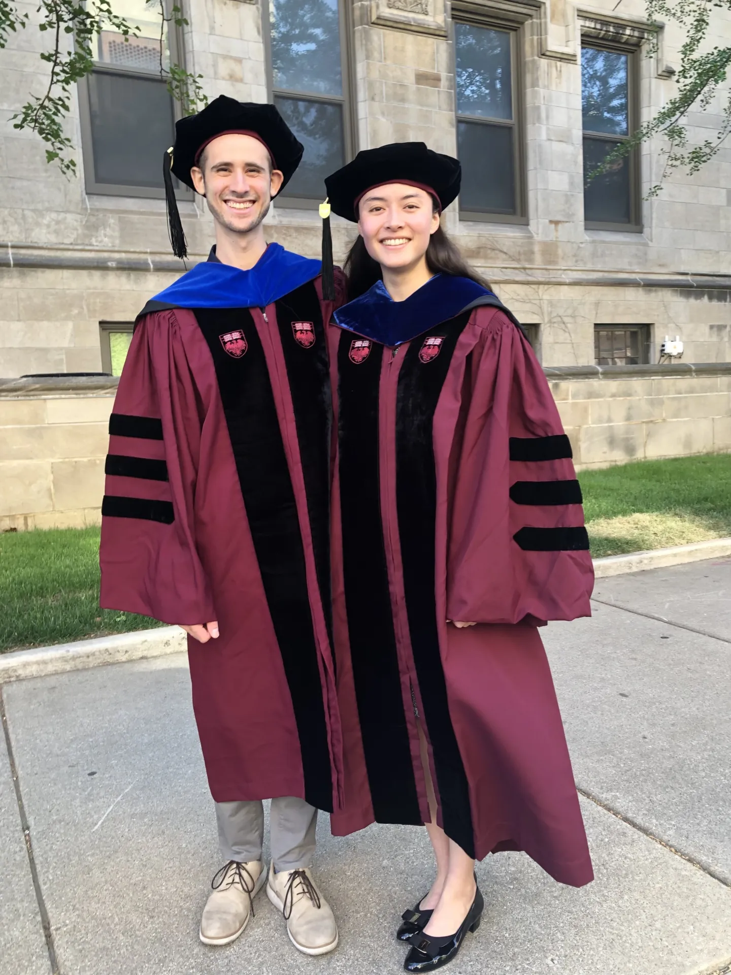 Two doctoral graduates in maroon academic regalia standing outside on the University of Chicago campus.