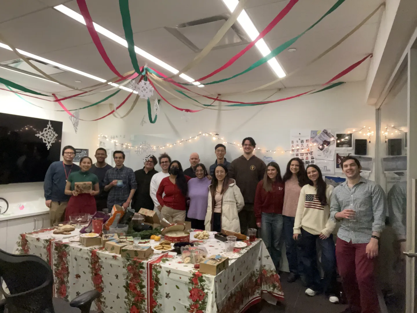 Group of students and researchers gathered around a holiday party table decorated with festive streamers, lights, and food in a laboratory lounge.