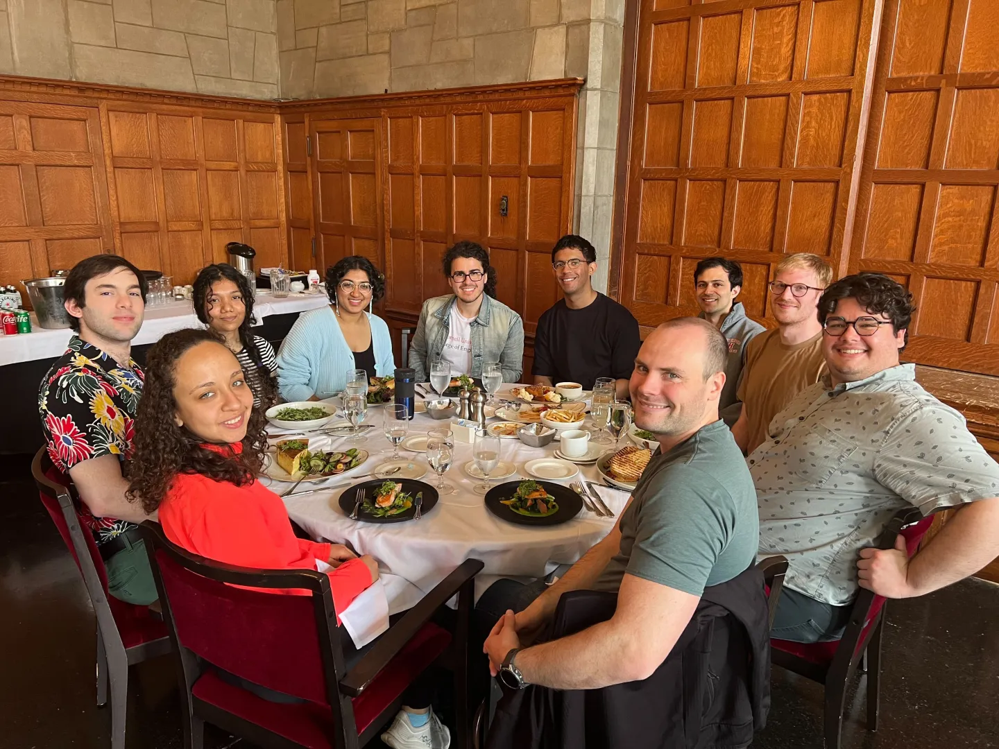 Graduate students and researchers seated around a dining table during a group lunch in a wood-paneled university hall.