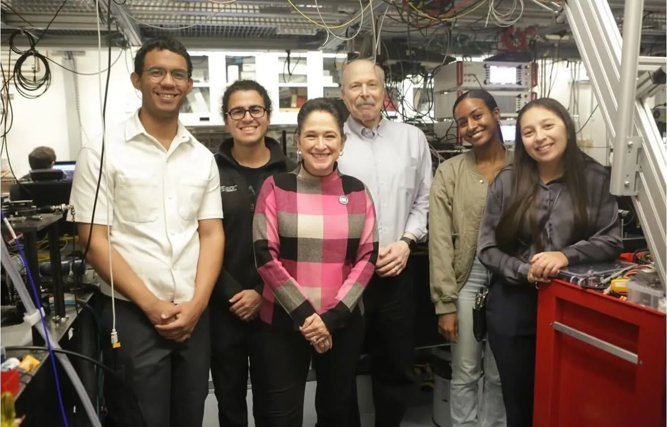 Researchers and visitors posing together inside a nanotechnology laboratory filled with scientific equipment and overhead wiring.