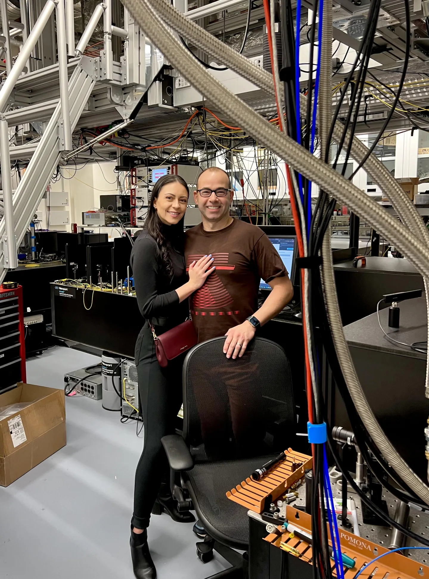 Couple posing together inside a nanotechnology laboratory surrounded by scientific instruments and wiring.