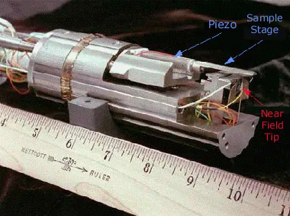 Photograph of a near-field scanning optical microscopy (NSOM) experimental setup showing the near-field probe tip, piezo positioning system, and sample stage. A ruler provides scale for the microscope assembly used in nanoscale optical measurements.