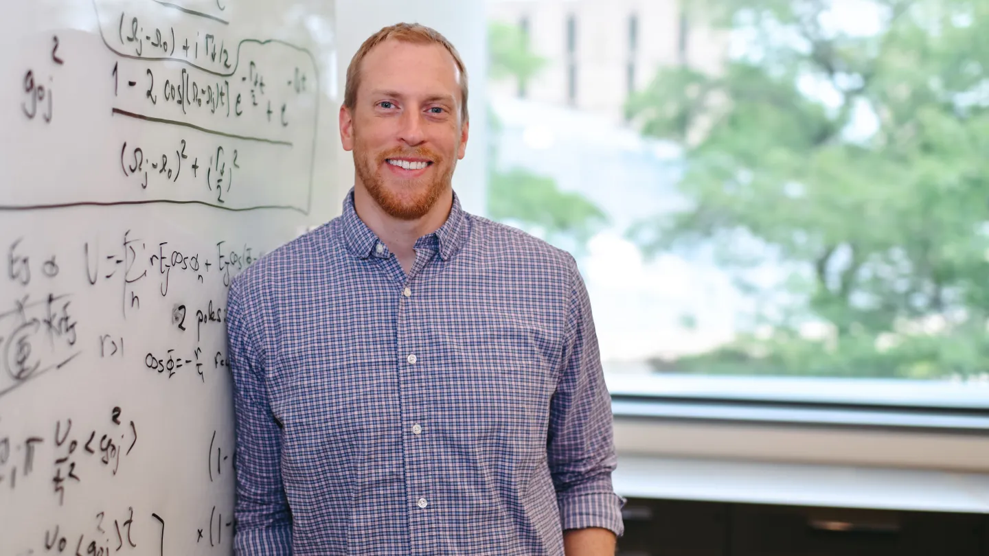 Man in button-down shirt next to whiteboard with physics equations