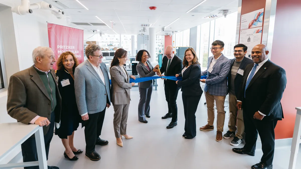 A group of nine people in professional attire celebrate a ribbon-cutting ceremony in a brightly lit modern laboratory. Two individuals in the center are cutting a blue ribbon.